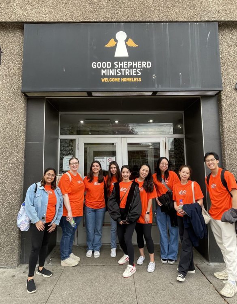 The Dream Marketing team wears orange t-shirts, and pose for a photo after a volunteering day, in front of doors for the charity, Good shepherd Ministries