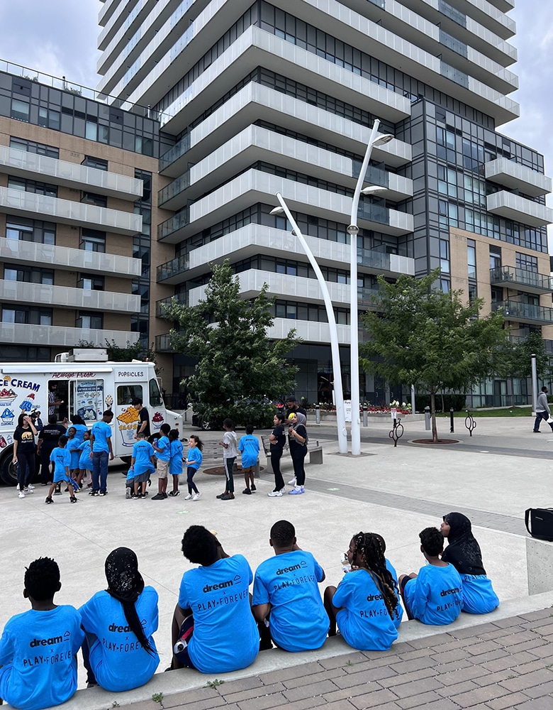 Group of children wearing matching blue "Dream, Play Forever" T-shirts gathered in a concrete city park, some sitting on steps while others crowd around a vibrant ice cream truck, surrounded by tall urban high-rise buildings.
