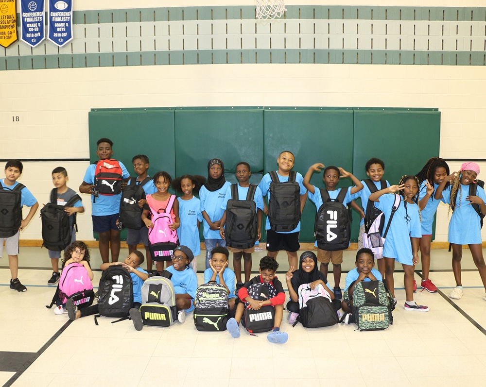 A large group of kids posing for a photo in a gymnasium wearing blue shirts and backpacks for the Dream Community Foundation