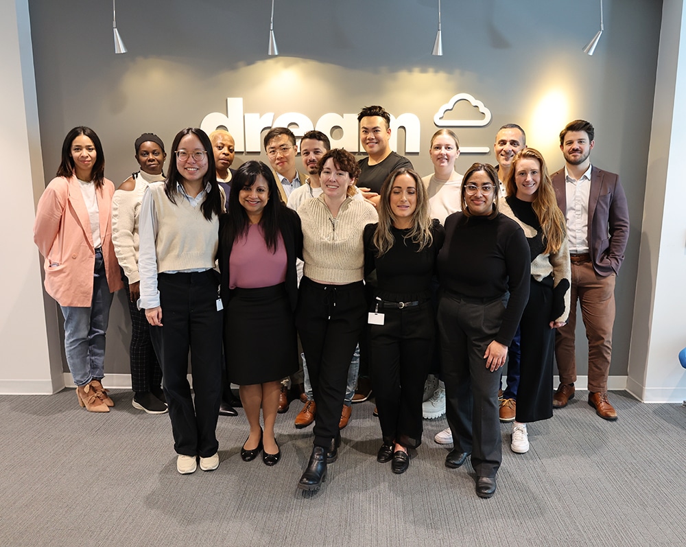 Diverse group of team members from the Diversity, inclusion and advancement group wearing business casual outfits, standing Infront of a grey wall that reads Dream with a cloud and line underneath