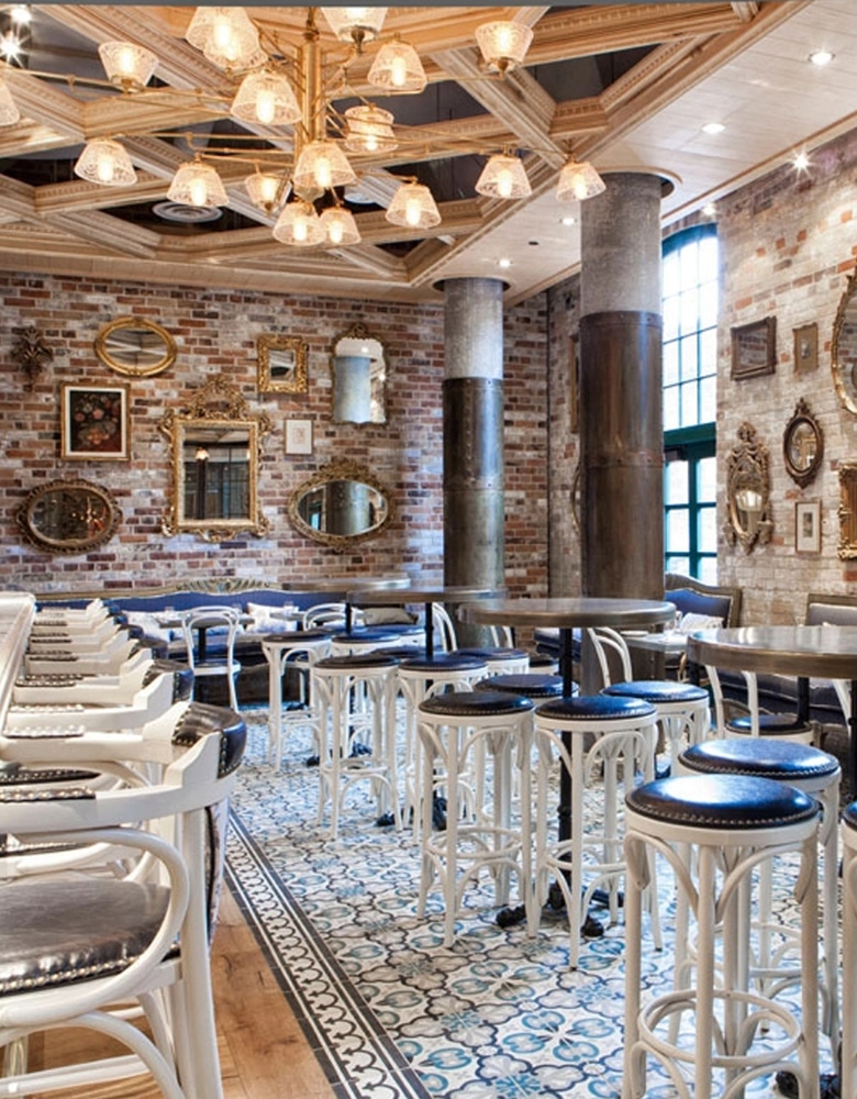 Interior of Cluny Bistro at the Distillery featuring vintage-modern decor, patterned tile flooring, exposed brick walls, white bar stools, round tables, ornate chandelier, and industrial columns.
