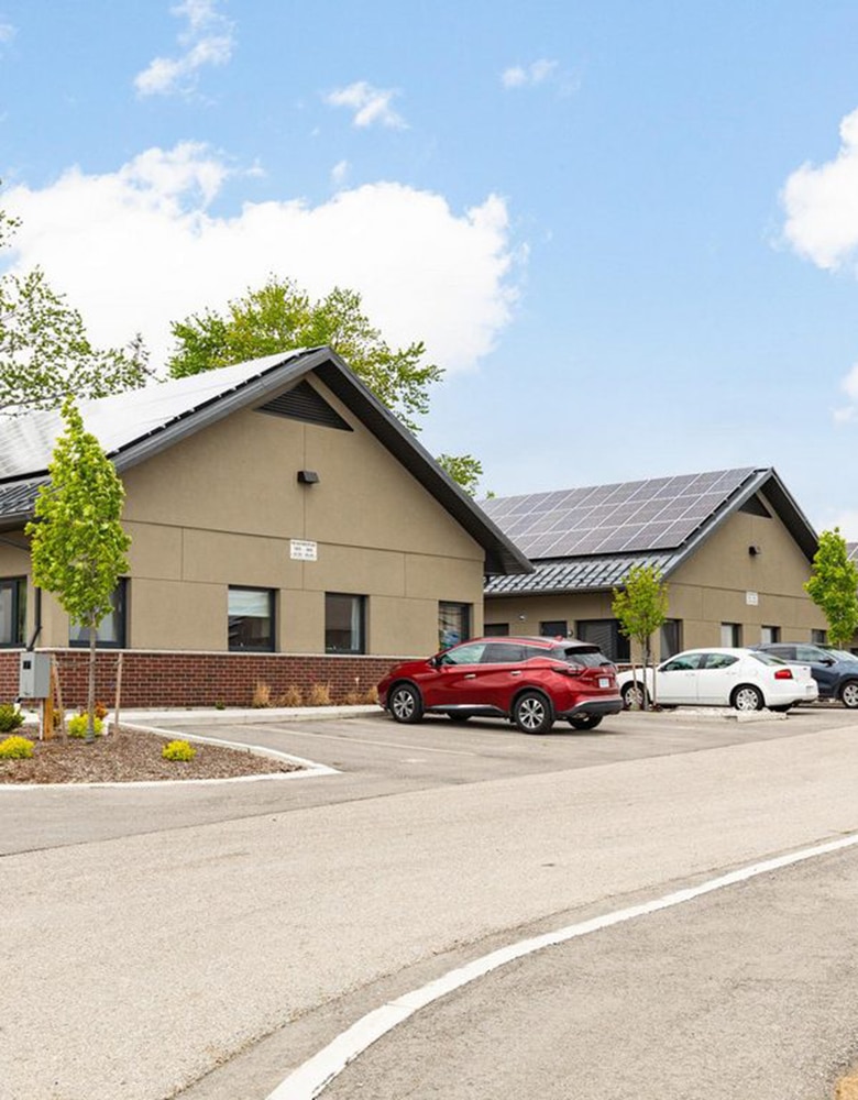 Parking lot with three cars in front of a beige building featuring solar panels on the roof, brick accents, windows, and surrounding trees under a partly cloudy sky