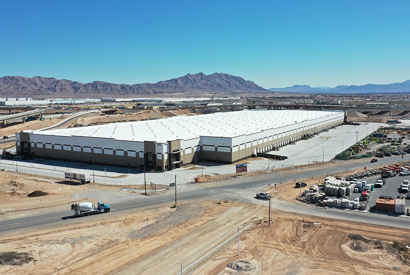 Large industrial warehouse with a white roof and beige walls in a desert area, surrounded by construction vehicles, materials, and mountains under a clear sky