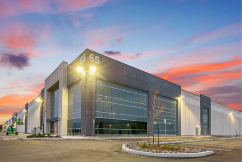 Modern industrial building with large glass windows and the number "65" displayed on the corner, surrounded by small trees and a paved area under a colorful sunrise or sunset sky