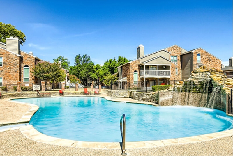 Outdoor swimming pool with rippling water and a stone deck, surrounded by two-story brick apartment buildings with balconies; a pergola stands to the left and a picnic table with an orange umbrella sits near the center, under a clear blue sky