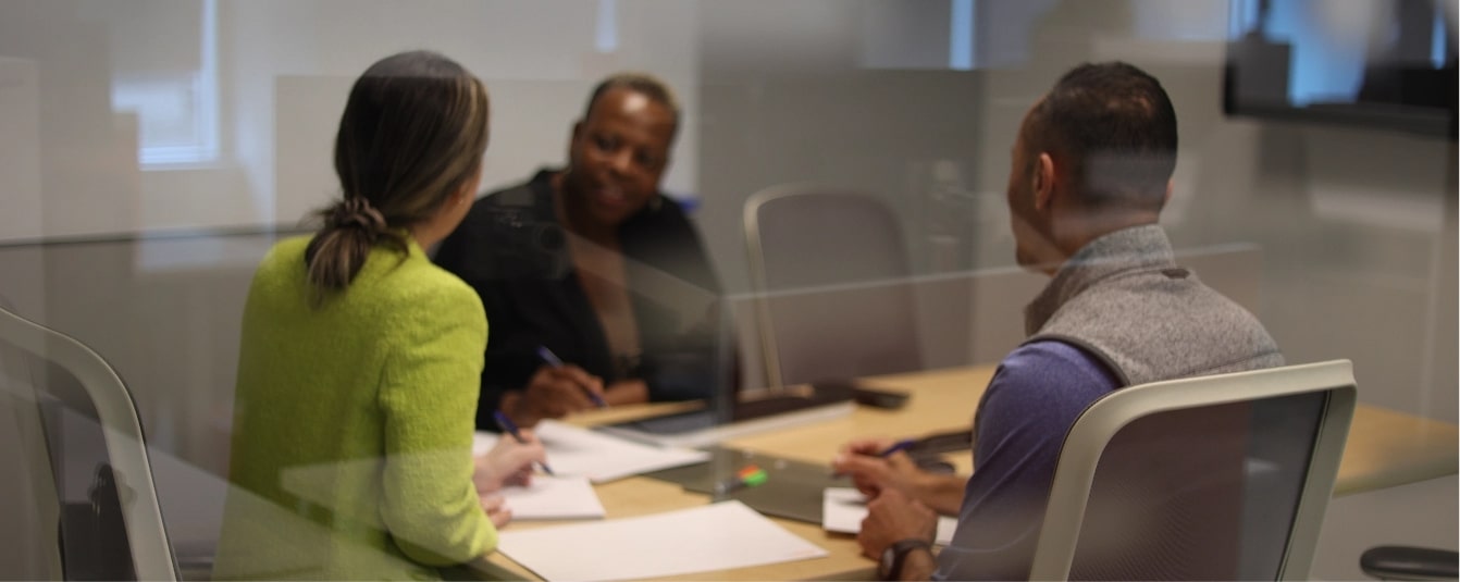 three people, two women and man in a boardroom taking notes while discussing a meeting