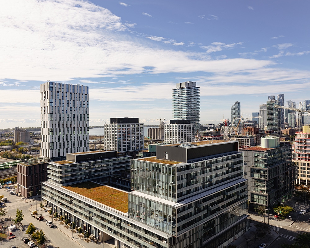 Cityscape featuring a multi-story building with a green roof in the foreground, surrounded by modern high-rise buildings, including a tall white patterned tower and a glass skyscraper, with cranes and scattered clouds in the background