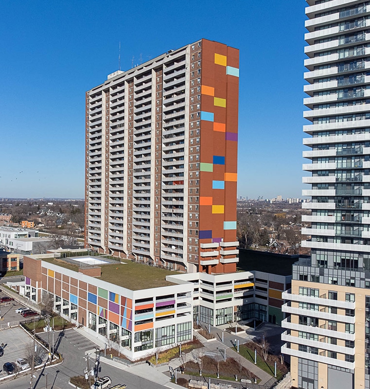 Urban scene featuring two tall buildings at Weston Commons, one with a colorful facade of orange, red, blue, and yellow panels and balconies, and the other with a sleek modern design of horizontal white lines and glass windows, with a parking lot and clear blue sky in the foreground.