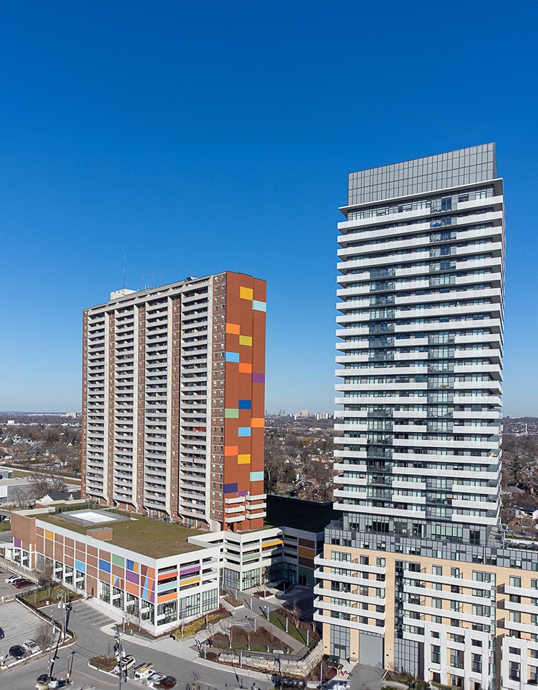 Urban scene featuring two tall buildings at Weston Commons, one with a colorful facade of orange, red, blue, and yellow panels and balconies, and the other with a sleek modern design of horizontal white lines and glass windows, with a parking lot and clear blue sky in the foreground.