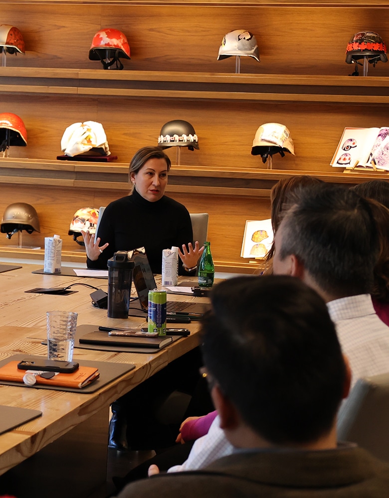Ana Bailao, speaking in a meeting room with decorated helmets displayed behind her, addressing an audience seated around a table with water bottles, notebooks, and laptops