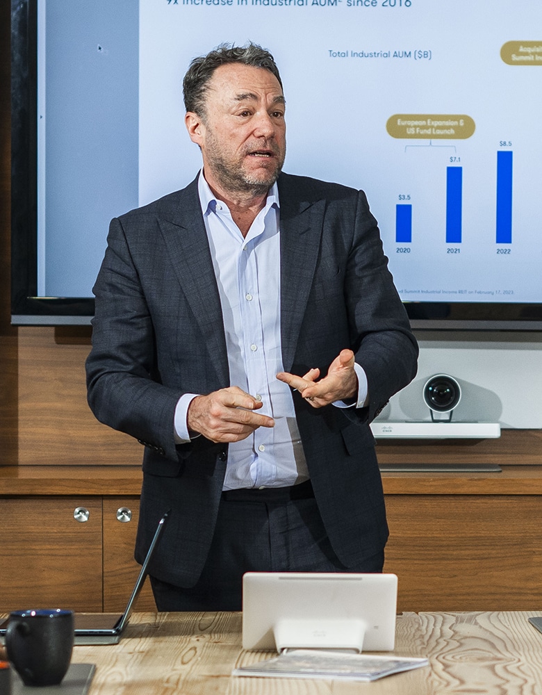 Michael Cooper, dressed in a shit, presenting in front of a screen displaying industrial AUM growth chart from 2020 to 2022, with key milestones including European expansion and Summit Industrial Income REIT acquisition