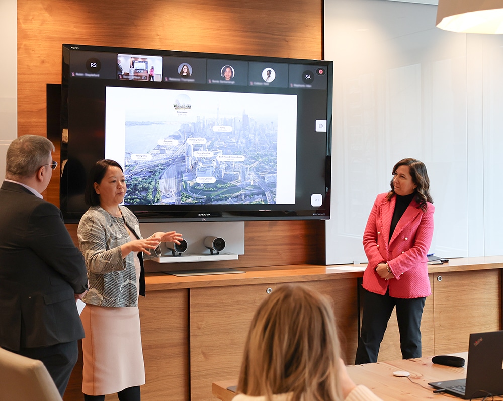 Tsering Yangki, Tony Medieros and Anna Bailao, in a meeting room with one person presenting an aerial city map on a large screen, featuring labeled locations and video call participants