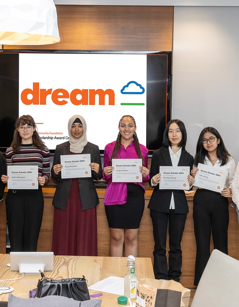 Four women stand shoulder to shoulder in a boardroom holding their certificates during the Scholarship Award Ceremony, in front of a screen that reads "Dream"