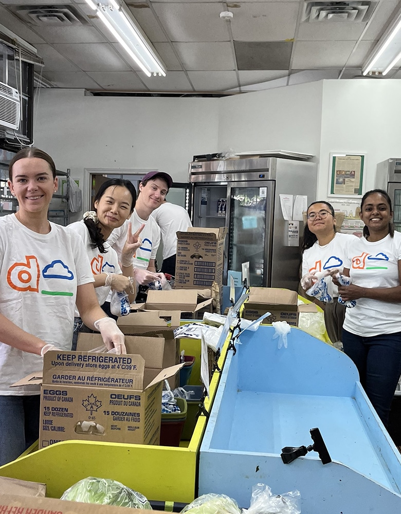 Group of people in a food pantry or distribution center wearing white t-shirts with an orange "d" and blue cloud logo, standing behind tables filled with boxes, eggs, and cabbages, with refrigerators and shelves in the background
