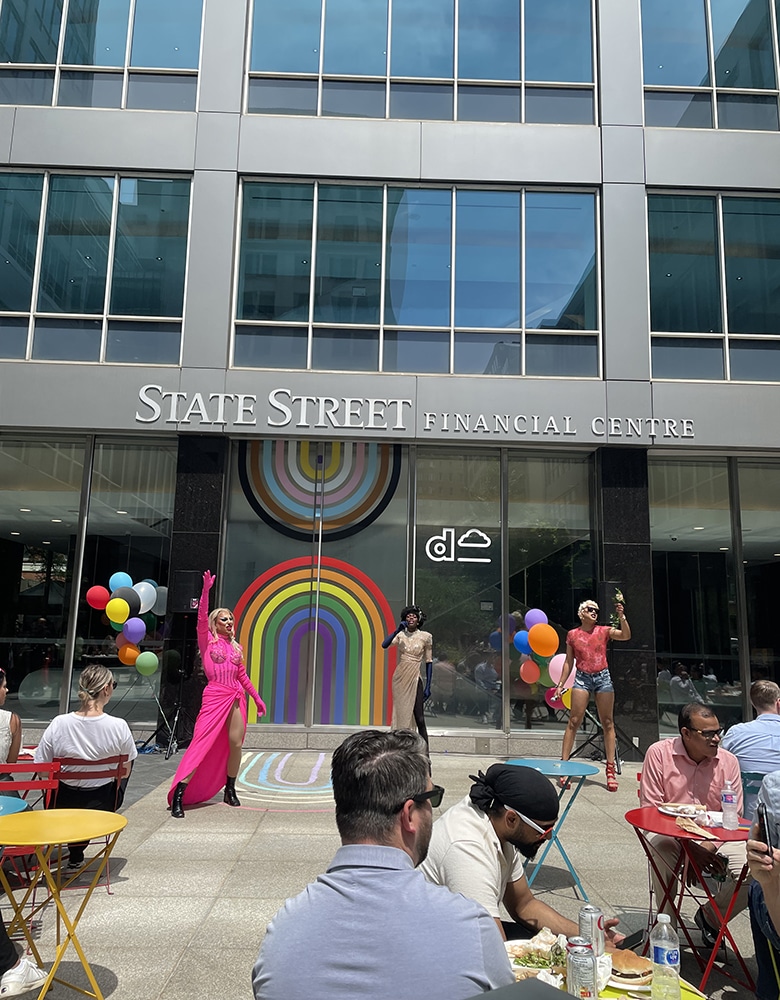 Pride event in front of the courtyard at State Street Financial Centre featuring three drag performers in colorful outfits on a stage with a rainbow backdrop and balloon decorations, with attendees seated at tables watching the show
