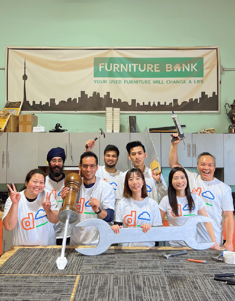 Group of people wearing matching white t-shirts with colorful logos, standing in front of a banner that reads "FURNITURE BANK YOUR USED FURNITURE WILL CHANGE A LIFE," holding tools and objects including a wrench, hammer, and giant pepper grinder, with cabinets and stacked cups in the background