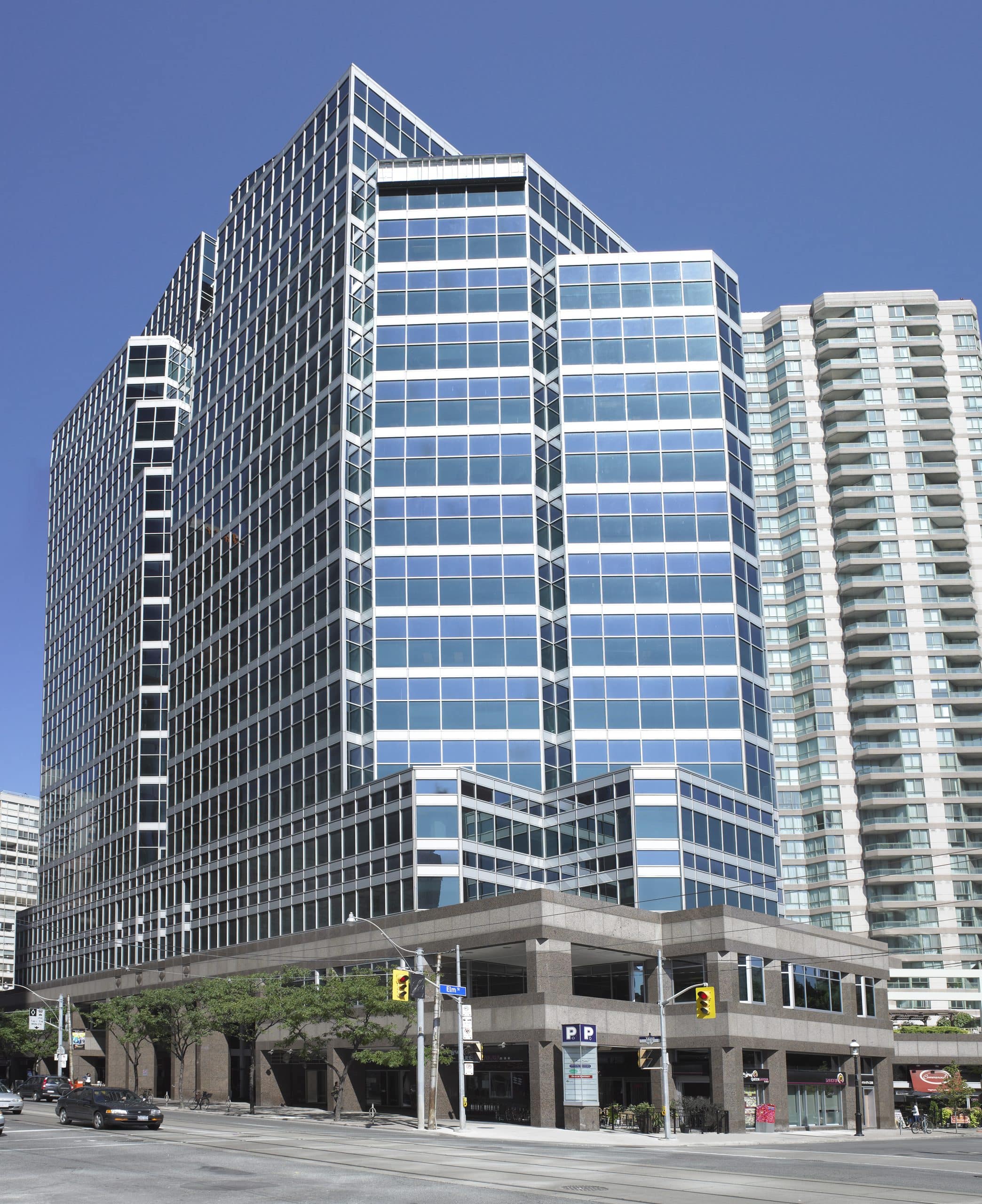 Tall modern office building at 655 Bay STreet, in Toronto, with glass exterior reflecting the blue sky, located at a street intersection with traffic lights, street signs, and parked cars; a second high-rise building is visible in the background