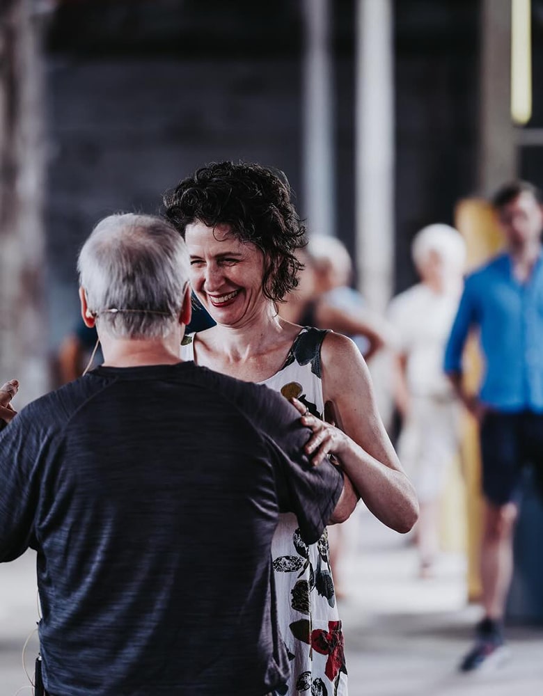 Two people dancing closely, one with short gray hair in a dark shirt and the other with curly dark hair in a sleeveless floral dress, with others in the background observing or participating