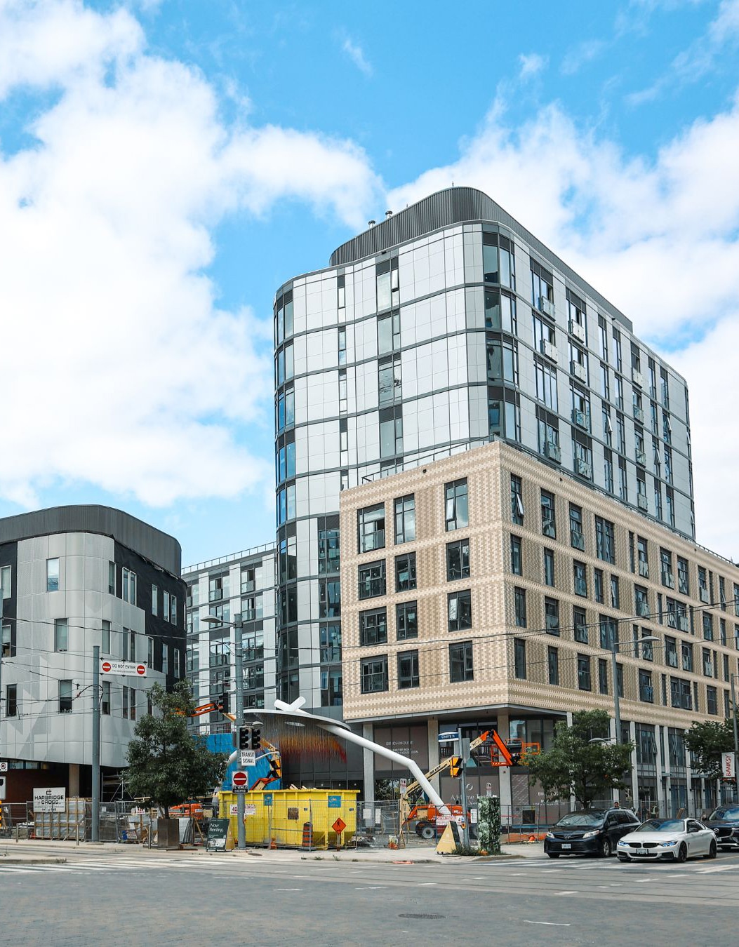 Modern urban scene with multi-story buildings featuring sleek designs and large glass windows, construction equipment and barriers at street level, and a few parked cars under a partly cloudy sky