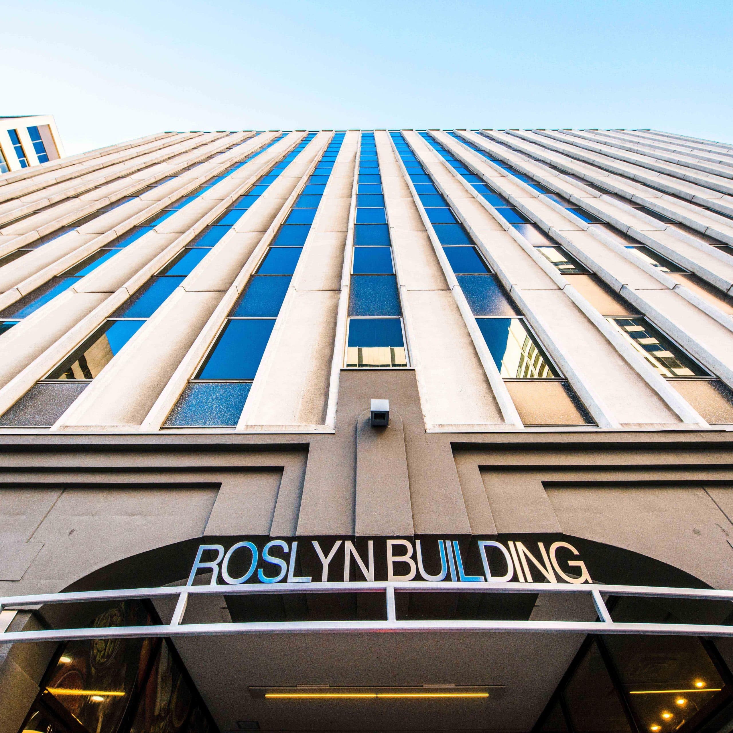 Upward view of the Roslyn Building, a tall modern structure with multiple floors and large vertical windows reflecting the blue sky