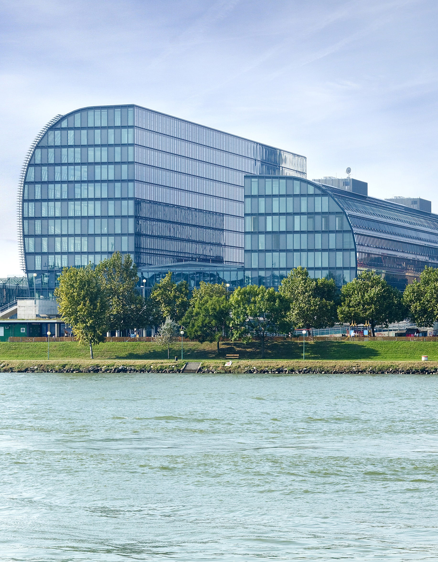 Modern glass building with a curved design on the left side, located near a body of water with trees and greenery in the foreground, under a clear sky with light clouds