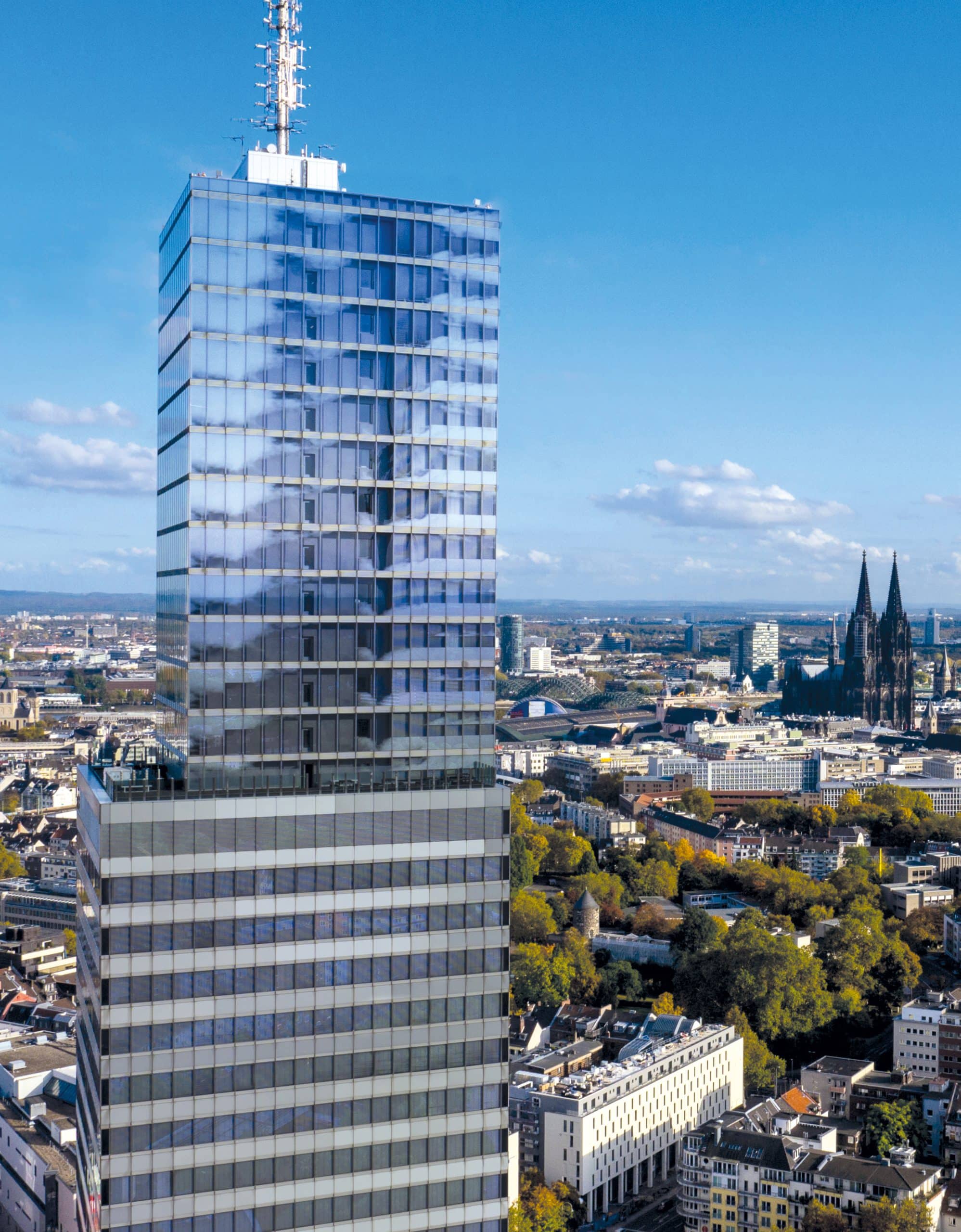 Tall modern skyscraper with reflective glass exterior and rooftop antennas, set against a cityscape featuring various buildings and a cathedral with twin spires under a partly cloudy sky