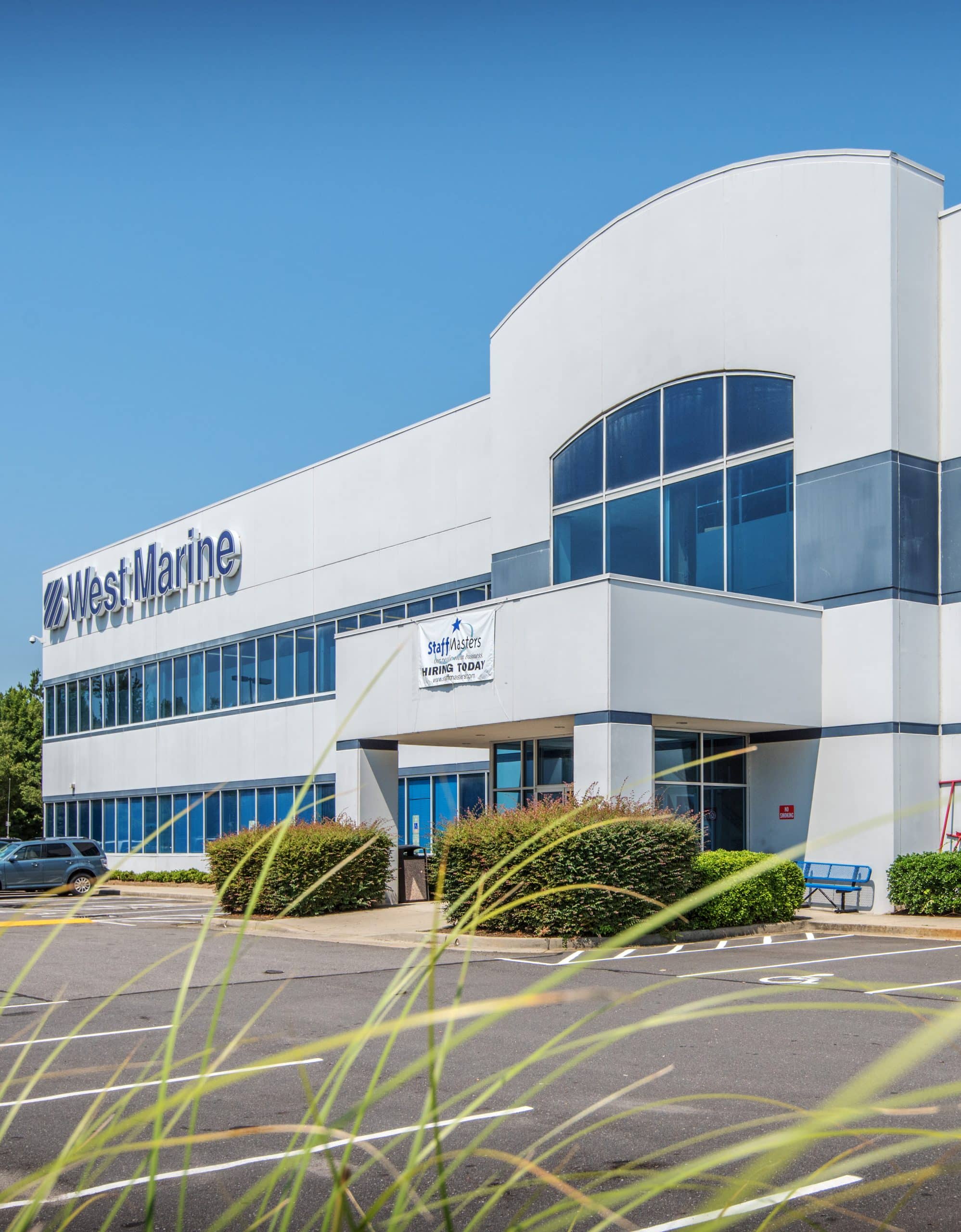 Modern commercial building with a "West Marine" sign and large windows; a "StaffMasters Hiring Today" banner is displayed, with greenery and an empty parking lot in the foreground