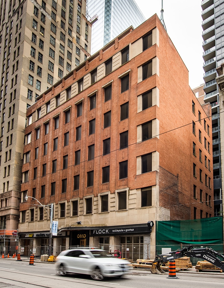 Multi-story brick building with large rectangular windows and ground-level storefronts labeled "OMD" and "FLOCK rotisserie + greens"; construction equipment and green netting are visible on the right side, with a white car driving past on the urban street