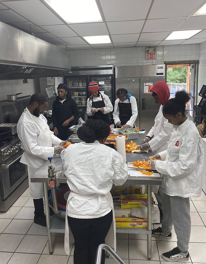 Seven People in chefs coats, standing in a commercial kitchen around steel table, prep. orange peppers