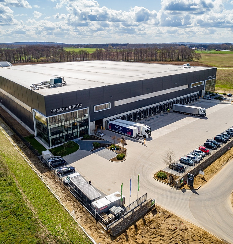 Large industrial warehouse labeled 'TIEMEX & STEFCO' with multiple loading docks, several trucks parked in front, a parking lot with cars, surrounding greenery, and open fields under a partly cloudy sky