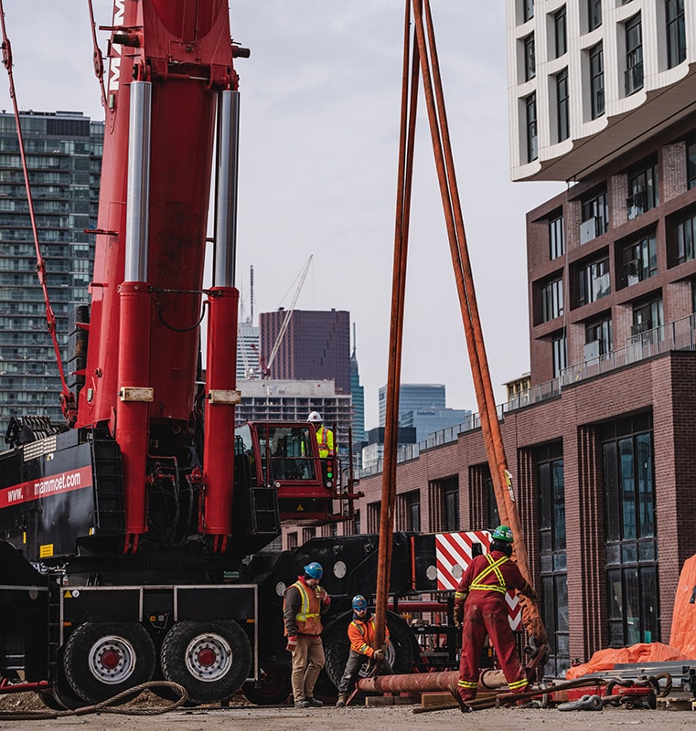 Construction site in the Canary District featuring a large red crane labeled 'www.mammoet.com', several workers in safety gear, and modern buildings and skyscrapers in the urban background