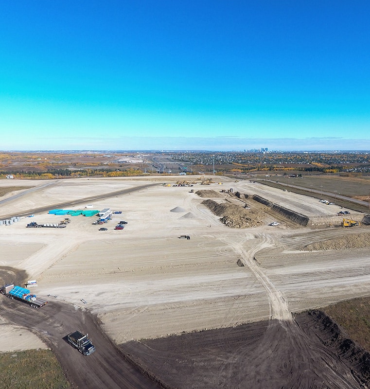 Large construction site with vehicles, equipment, dirt roads, trenches, and piles of earth, set against a rural or suburban landscape with a city skyline visible on the horizon under a clear blue sky