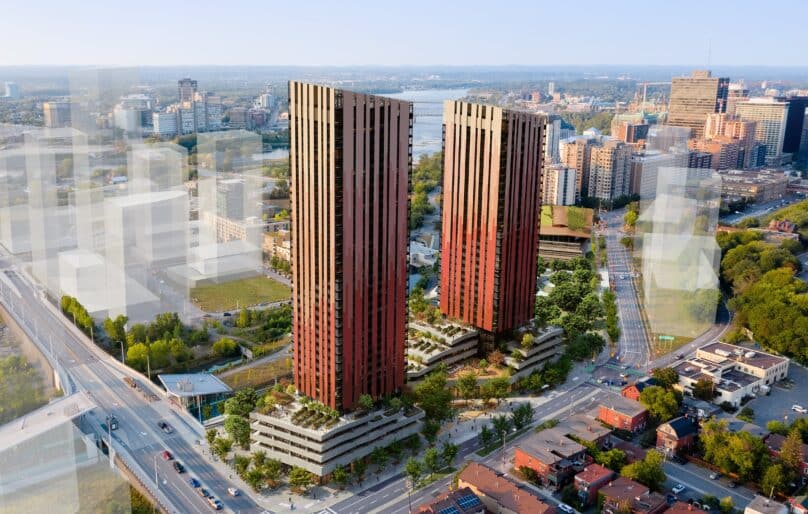 Aerial view of the Odenak building in Ottawa, showing two tall reddish-brown skyscrapers surrounded by greenery, nearby buildings, a major road with traffic, and a cityscape with high-rises and a river in the background