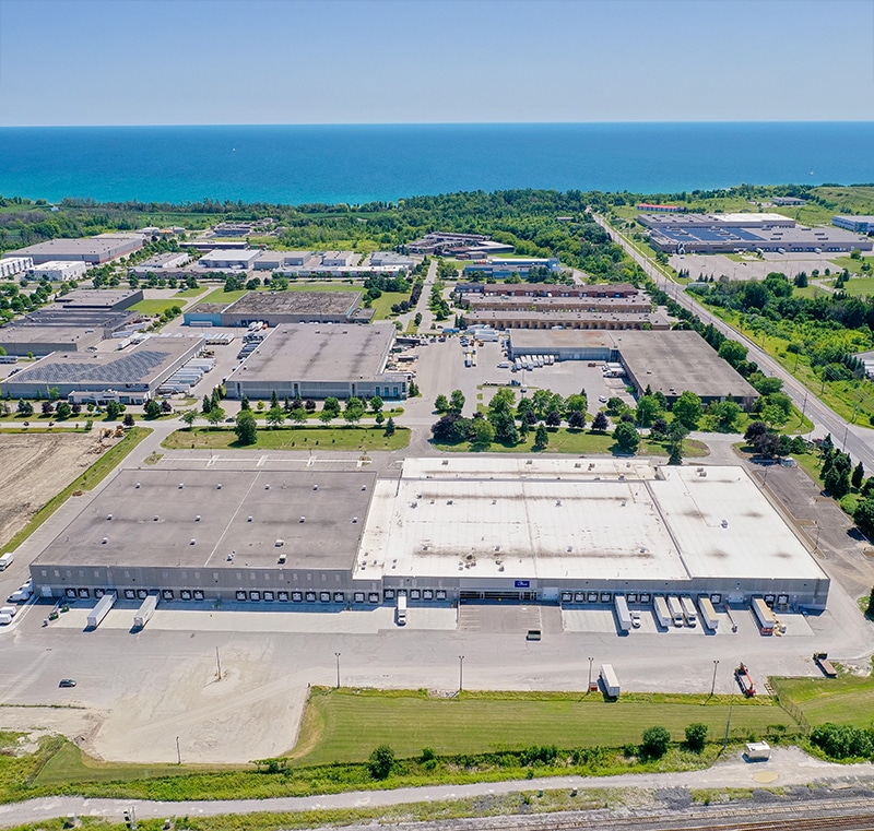 Aerial view of an industrial area near a large body of water, featuring a large white and cement toned warehouse with multiple loading docks and parked trucks. Surrounding the warehouse are other industrial buildings, parking lots, and green spaces with trees, with additional buildings and greenery extending toward the shoreline