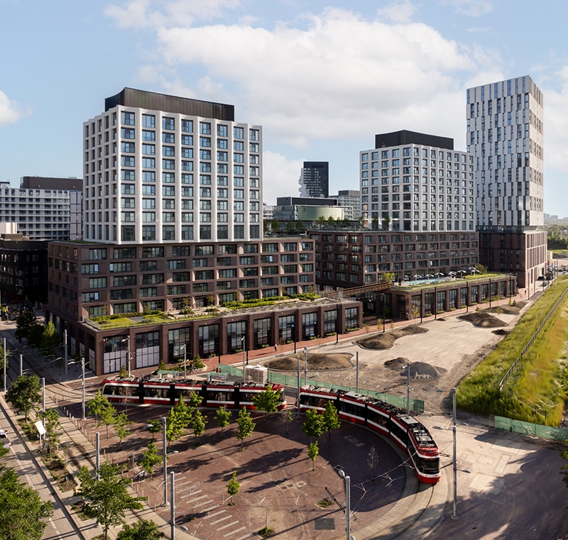 Modern urban area with multi-story buildings of varied architectural styles, including large windows and intricate designs. A TTC Street car, a red and white tram, travels along curved tracks in the foreground, surrounded by trees, streetlights, green spaces, and pedestrian pathways