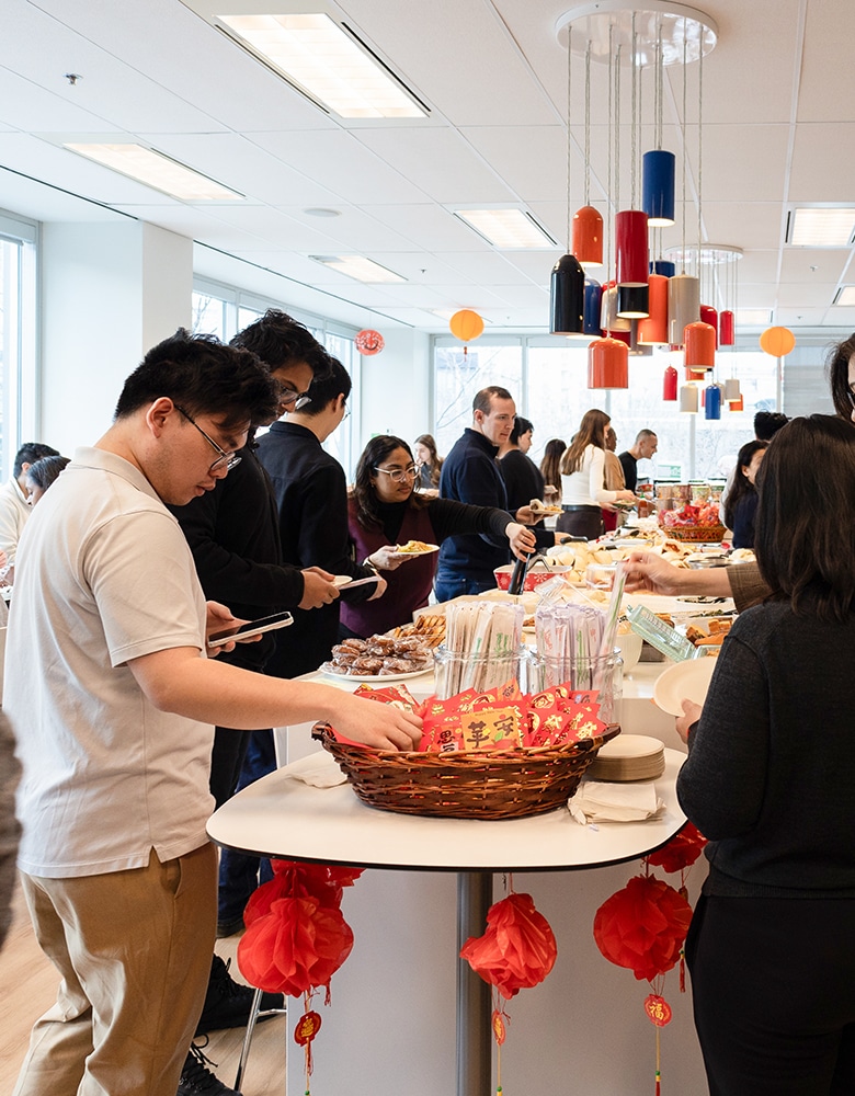 Lunar New Year celebration with people gathered around tables selecting traditional items; red lanterns hang overhead and red envelopes are prominently displayed, creating a festive and cultural atmosphere
