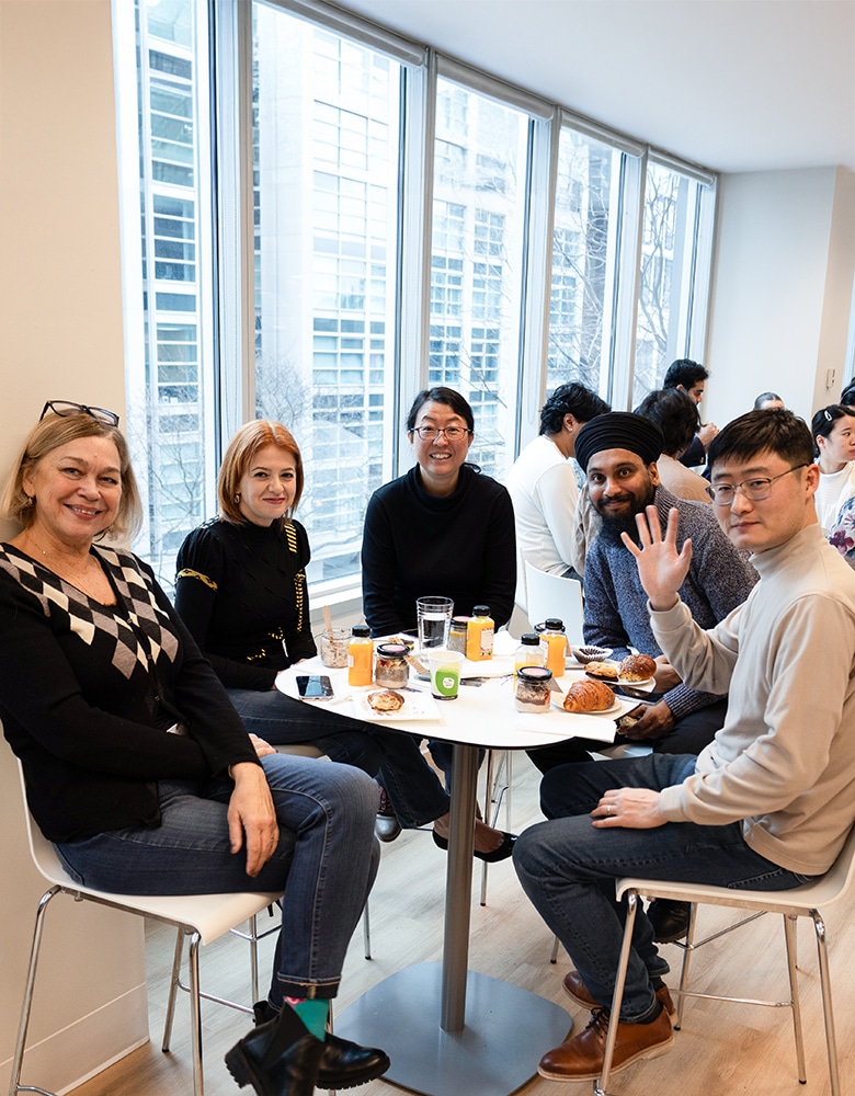 Group of people gathered around a table with assorted beverages and pastries, smiling and posing for a photo in a casual, social setting