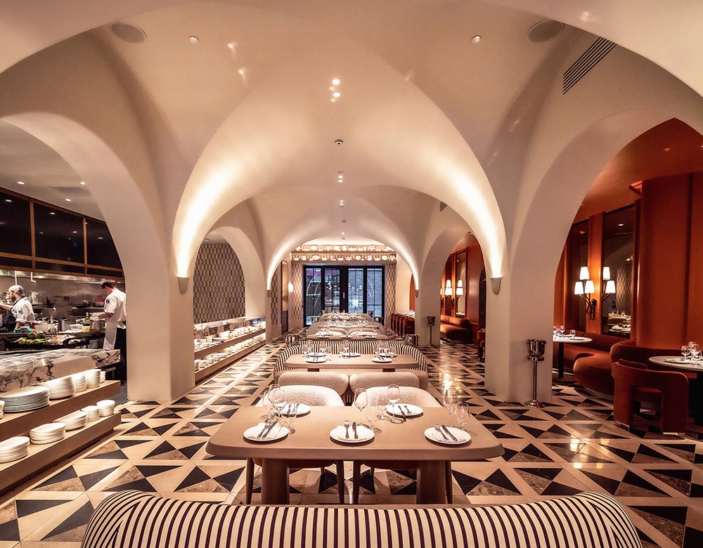 Interior of Daphne Restaurant showcasing a modern luxury space with warm inviting tones, perfectly centered tables and tableware under vaulted ceilings, sherpa-style chairs, zebra-striped couches, and floor tiles with triangle motifs