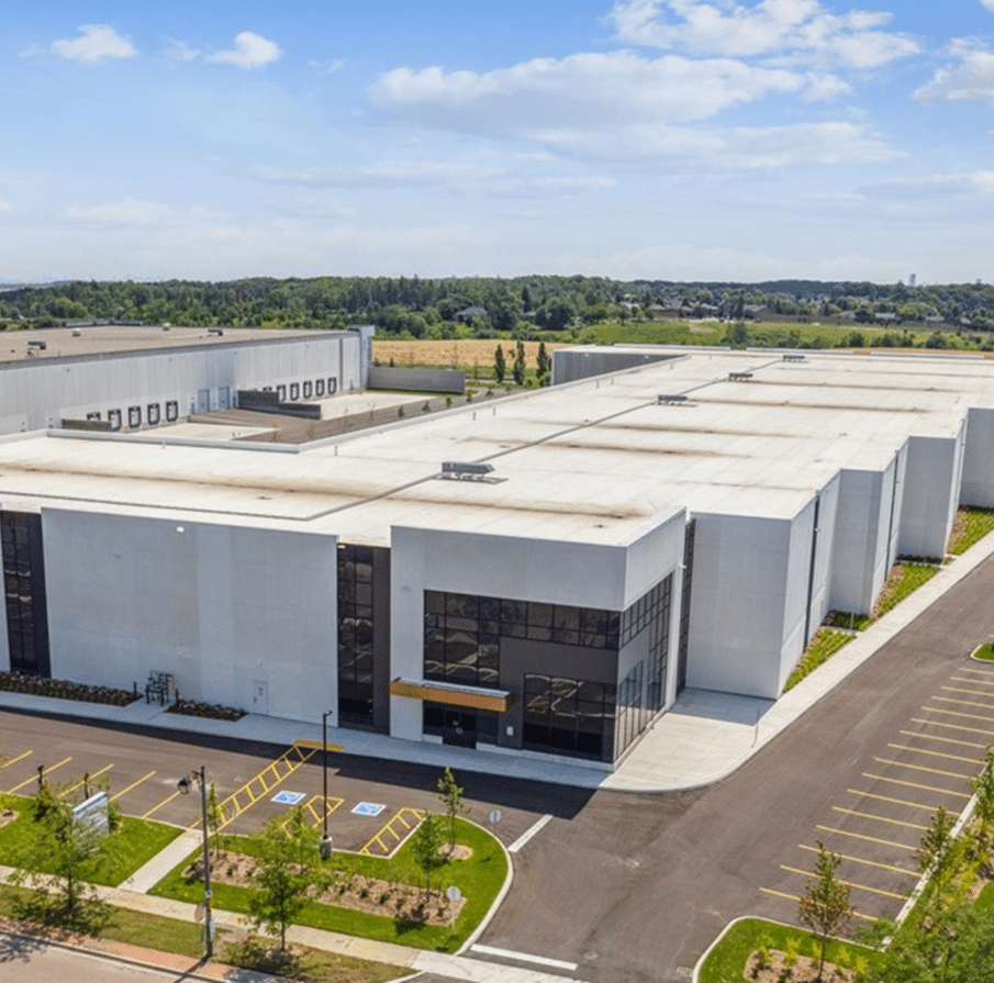 Modern white industrial building with a flat roof and clean, straight architectural lines; surrounded by lush green grass and trees, with an empty parking lot in the foreground